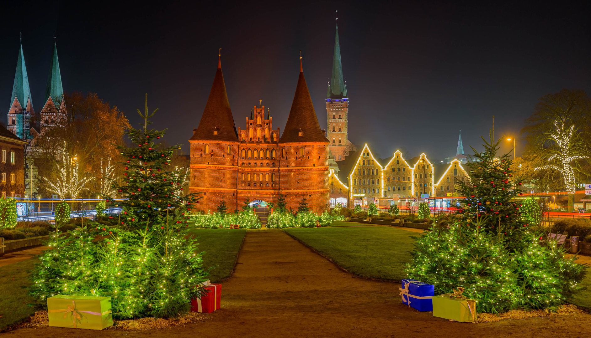 Das festlich beleuchtete Holstentor. Im Vordergrund geschmückte, erleuchtete Weihnachtsbäume, im Hintergrund die Salzspeicher.