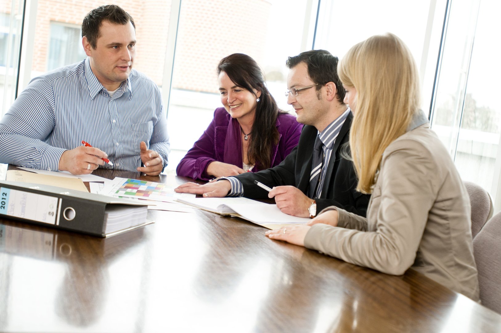 Eine Teambesprechung mit vier Personen (zwei Frauen, zwei Männer). Auf dem Tisch liegt ein Aktenordner. Eine Teambesprechung mit vier Personen (zwei Frauen, zwei Männer). Auf dem Tisch liegt ein Aktenordner.