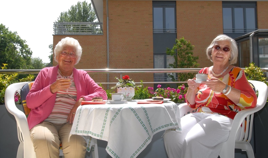 Zwei alte Damen auf einem Balkon. Sie trinken Kaffee und lächeln.