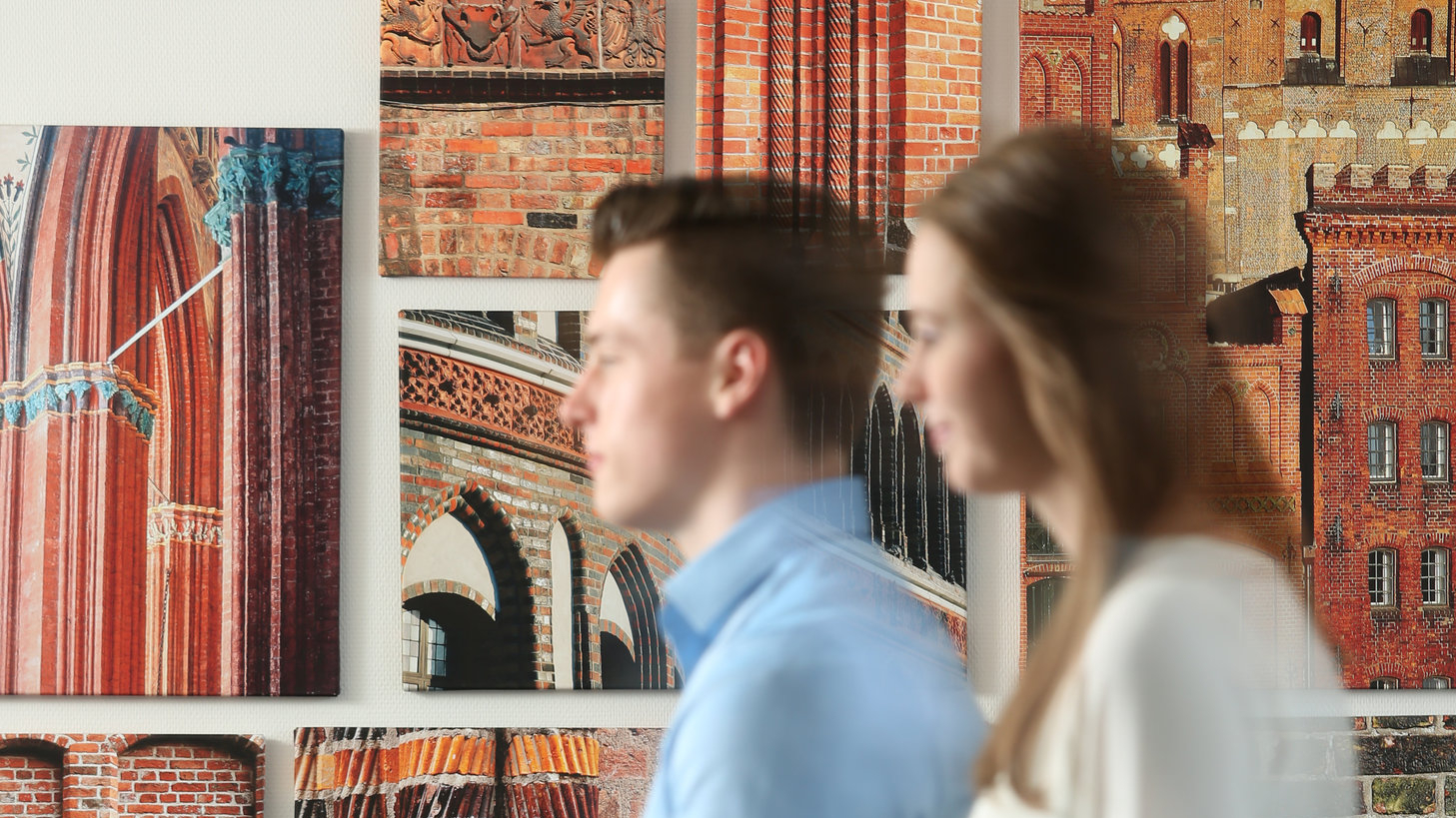 Zwei junge Menschen gehen an einer Wand vorbei. An der Wand sind Bilder mit Backsteinmotiven aus Lübeck zu sehen.