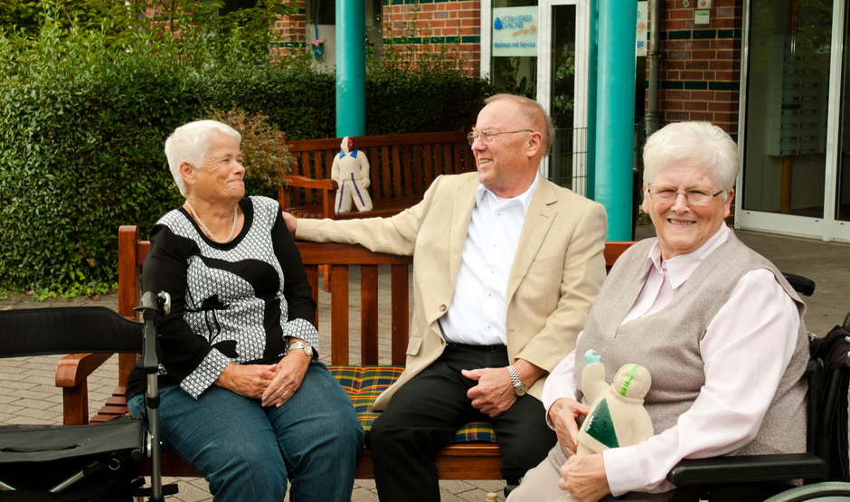Zwei gut gelaunte Damen und ein Herr sitzen auf einer Bank vor der Seniorenwohnanlage im Eulenspiegelweg.