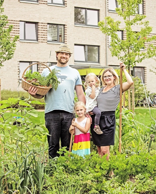 Nachbarschaftsgarten Vater, Mutter und zwei kleine Mädchen stehen mit einem Korb mit geerntetem Gemüse und einer Harke im Nachbarschaftsgarten. Im Hintergrund ein modernes Mehrfamilienhaus.