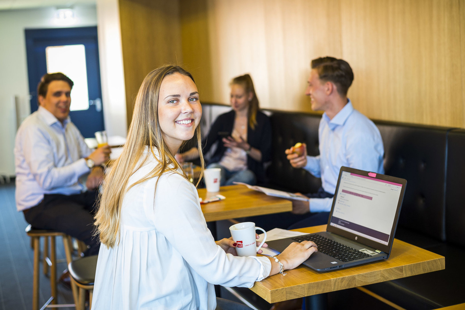 Vier Auszubildende der TRAVE an Bistrotischen. Im Vordergrund lächelt eine junge Frau (Auszubildende) mit Laptop in die Kamera. Vier Auszubildende der TRAVE an Bistrotischen. Im Vordergrund lächelt eine junge Frau (Auszubildende) mit Laptop in die Kamera.