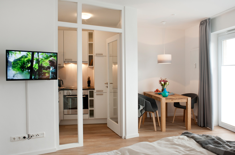 Blick in den Raum: Flachbildfernseher an der Wand, rechts Esstisch mit zwei Stühlen, dazischen der Blick in die Küche.